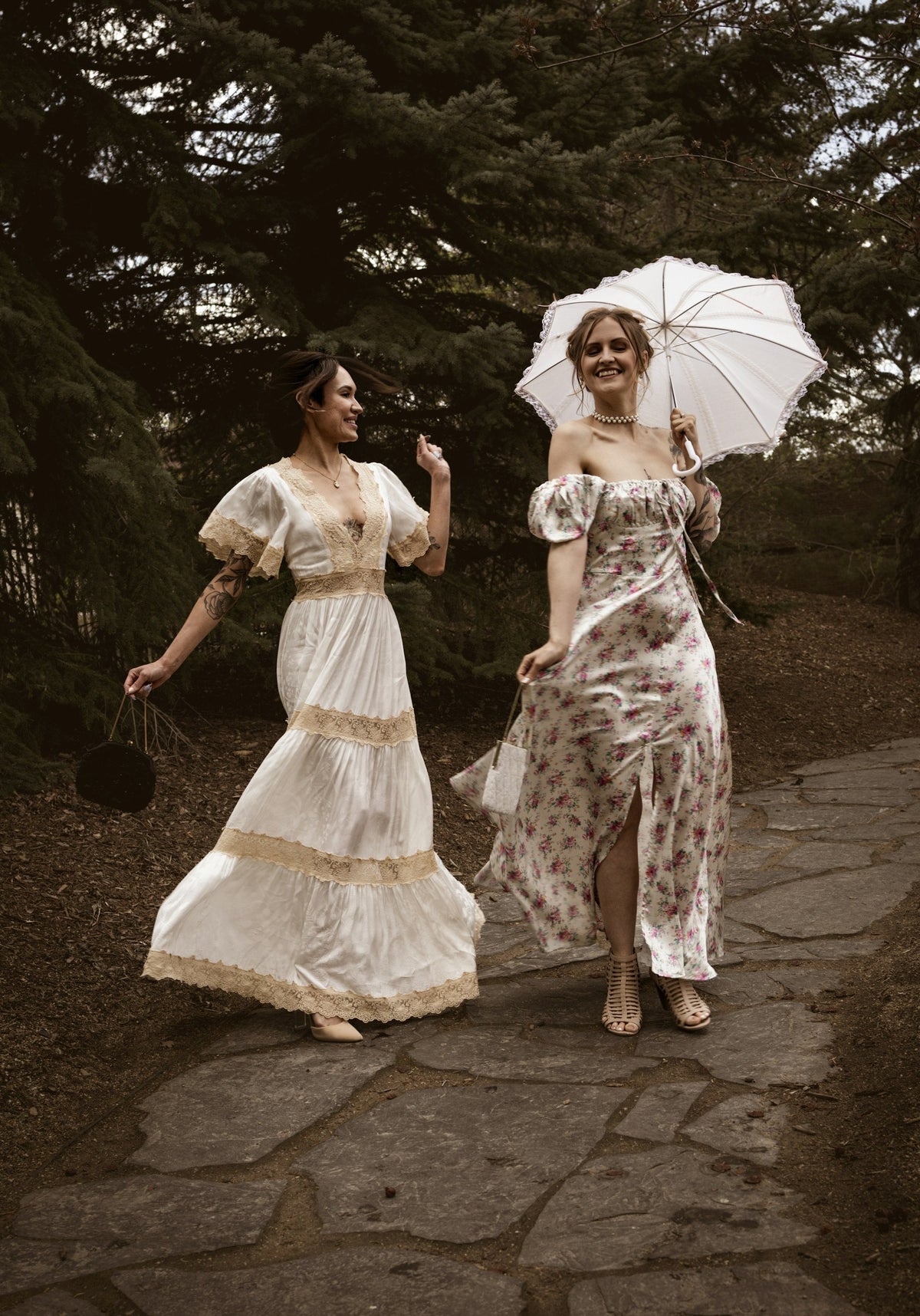 Two women in vintage dresses, one holding a white lace parasol, walking along a garden path
